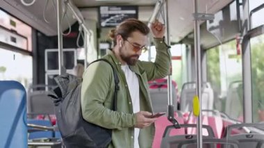 Sırt çantası ve güneş gözlüğü takan, otobüsle giden, ayakta duran meşgul bir adamın yan görüntüsü. Sakallı, kulaklık takan, akıllı telefon tutan yakışıklı bir erkek. Toplu taşıma kavramı.
