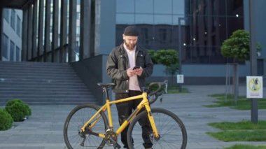 Modern binanın yanında duran sarı bisikletli genç adamın ön görüntüsü. Sakallı, sıradan kıyafetler giyen, akıllı telefon kullanan, kitap okuyan yakışıklı bir erkek. Kentsel yaşam tarzı kavramı.