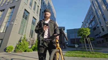 Bisikletle dışarıda duran sakallı, yakışıklı genç bir adam. Teslimatçı, kurye günlük kıyafetler giyiyor, akıllı telefon kullanıyor, mesaj yazıyor. Kentsel yaşam tarzı kavramı.