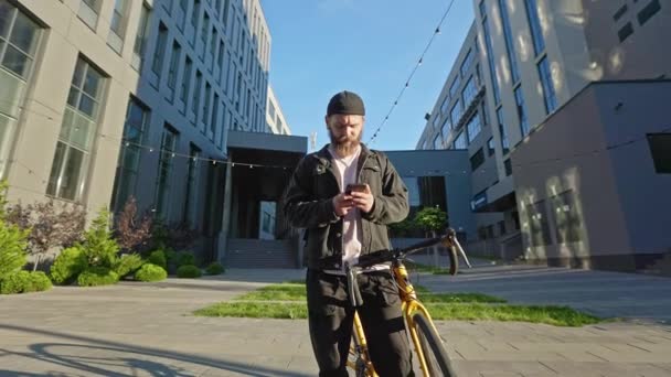 年轻男子骑着自行车 拿着智能手机 在前面看 留着胡子 穿着休闲装 发短信 发短信的男人 城市生活方式的概念 — 图库视频影像
