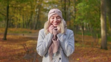 Front view of smiling, young lady walking in park in autumn. Attractive, blonde woman standing, hugging, looking at camera, smiling, enjoying. Concept of happiness and coziness.