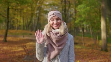 Front view of sincere, young lady standing in park, waving hello. Beautiful, blonde female looking at camera, smiling, wearing stylish clothes. Concept of happiness and coziness.