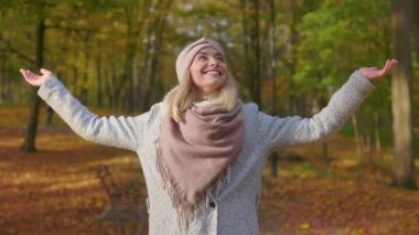 Front view of beautiful, blonde woman walking in park, wearing stylish clothes. Attractive, young female looking up, smiling, stretching hands. Concept of coziness and happiness.