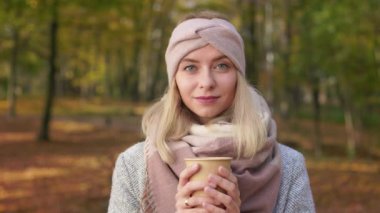 Front view of smiling blonde woman standing in park in autumn. Pretty, young lady holding paper cup with coffee, enjoying, looking at camera. Concept of happiness and coziness.