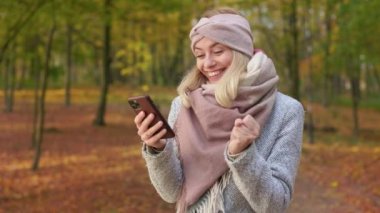 Side view of cheerful, young lady walking in park in autumn. Attractive, blonde woman using smartphone, texting, chatting, jumping, smiling, surprised. Concept of sincere emotions.