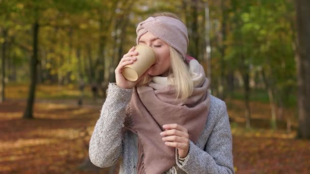 公園に立って幸せな 幸せな女性のフロントビューでは 紙コップを保持します 美しく ブロンドの女性は 目を閉じてコーヒーを飲み 楽しんでいます 秋と幸せの概念 — ストック動画