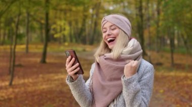 Side view of attractive, blonde woman walking in park in autumn. Beautiful, young female using smartphone, texting, showing hurray, smiling. Concept of sincere emotions.