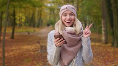 Front view of cheerful, young lady standing in park with golden trees. Beautiful, blonde woman using smartphone, chatting, looking at camera, smiling. Concept of autumn and fall.