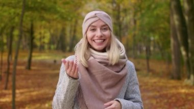 Front view of beautiful, blonde woman wearing stylish clothes, standing in park. Attractive, young female looking at camera, talking. Concept of happiness and sincere emotions.