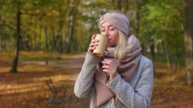 Front view of happy, glad woman walking in park in autumn. Attractive, blonde woman standing, drinking, sipping coffee, enjoying, smiling. Concept of happiness and coziness.