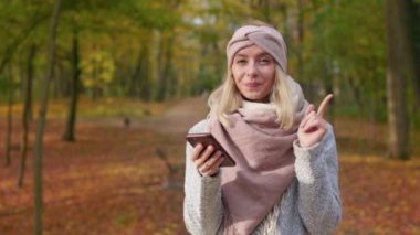 Front view of cheerful, young lady standing in park with golden trees. Attractive, blonde woman using smartphone, texting, chatting, thinking. Concept of modern lifestyle.