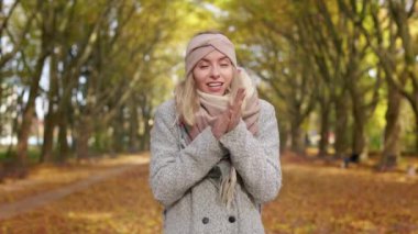 Front view of attractive, blonde female walking in park in cold weather. Beautiful, young lady standing, looking at camera, warming hands, smiling. Concept of autumn and fall.