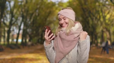 Side view of blonde woman standing in park in autumn. Attractive, young female holding, using smartphone, texting, messaging, glad, happy. Concept of happiness and coziness.