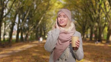 Front view of glad, young female walking in park, standing. Attractive, blonde female talking on smartphone, smiling, holding paper cup, drinking coffee. Concept of modern lifestyle.