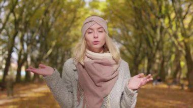 Front view of beautiful, young female standing in park, in autumn. Attractive, blonde female walking in park, looking at camera, telling, explaining. Concept of modern lifestyle.