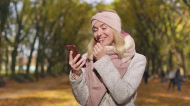 Front view of blonde woman walking in park in autumn. Young female standing, holding, using smartphone, texting, reading, happy, surprised. Concept of happiness and coziness.