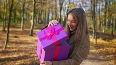 Side view of fashionable, pretty lady walking in park in autumn. Attractive, curious, young female standing, opening present, gift box, enjoying. Concept of happiness and joy.