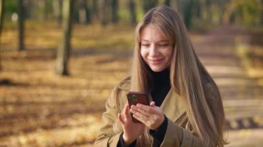 Sonbaharda parkta akıllı telefon kullanan güzel, genç bir bayanın yan görüntüsü. Şaşırmış, mutlu, mutlu kadın mesajlaşıyor, kayıyor, mesajlaşıyor, gülümsüyor. Mutluluk ve neşe kavramı.