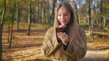 Mutlu, genç bir bayanın parkta yürümesini ön planda görmek. Akıllı telefon kullanan, kaydıran, mesaj atan, okuyan, gülümseyen, merak eden çekici bir kadın. Modern yaşam tarzı kavramı.