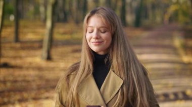 Güneşli sonbahar havasında parkta yürüyen çekici bir kadın. Güzel, genç bir bayan ayakta, hayranlık duyuyor, eğleniyor, gülümsüyor, akıllı telefon kullanıyor. Doğayla uyum kavramı.