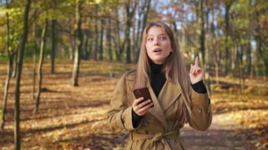 Parkta akıllı telefon kullanarak yürüyen güzel, genç bir kadının ön görüntüsü. Güzel, şık bayan mesajlaşması, mesajı, kaydırması, düşünmesi, hatırlaması. Modern teknoloji kavramı.