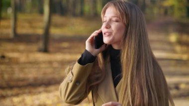 Sonbaharda parkta yürüyen mutlu, genç bir kadın. Güzel kıyafetler giyen, telefonda konuşan, gülümseyen çekici bir bayan. Doğanın güzelliği kavramı.