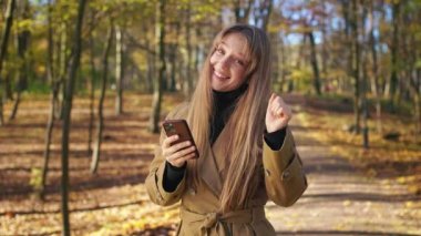 Parkta yürüyen, müzik dinleyen, dans eden pozitif, neşeli bayanın ön görüntüsü. Şık bir kadın şık bir siper giymiş, kameraya bakıyor. Modern yaşam tarzı kavramı.
