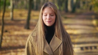 Front view of happy, elegant woman wearing fashionable clothes, walking in park. Beautiful, young lady standing, looking at camera, blinking. Concept of harmony with nature.