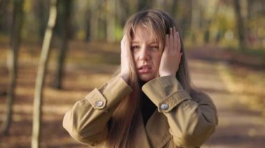 Side view of pretty, young female standing in park, having terrible headache. Tired, upset woman walking, touching, pressing head, closing eyes. Concept of modern lifestyle.