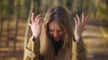 Front view of angry, young female standing in park, raising hands, quarreling. Pretty woman wearing fashionable clothes, looking at camera, upset. Concept of sincere emotions.