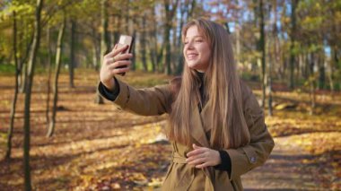 Side view of beautiful lady talking by video in park in fall. Beautiful, cheerful lady taking video, smiling, waving, showing, happy, glad. Concept of modern lifestyle.