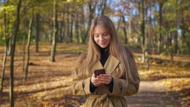 Front view of happy, cheerful woman enjoying sunny autumn weather in park. Attractive, young lady standing, using smartphone, closing eyes, smiling. Concept of beauty of nature.
