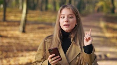 Front view of young, attractive woman standing in park, using smartphone. Beautiful, stylish female texting, messaging, thinking, remembering. Concept of modern urban lifestyle.
