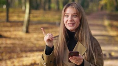 Front view of happy, cheerful lady walking in park, using smartphone. Attractive, young female standing, texting, scrolling, messaging, smiling. Concept of modern lifestyle.