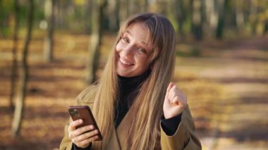 Front view of fashionable lady using smartphone, dancing, happy, glad. Cheerful, positive woman walking in park, standing, looking at camera, enjoying. Concept of happiness.