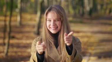 Front view of beautiful, young female walking in park in autumn. Attractive, cheerful lady standing, looking at camera, pointing by fingers, smiling. Concept of sincere emotions.