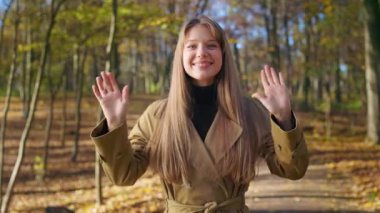 Front view of cute, positive lady walking, standing in park. Cheerful, young female looking at camera, smiling, waving, wearing stylish clothes. Concept of sincere emotions.