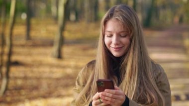 Parkta gezen, şık kıyafetler giyen mutlu, pozitif bir kadının yan görüntüsü. Çekici, genç bir bayan ayakta duruyor, akıllı telefon kullanıyor, kaydırıyor. Mutluluk ve keyif kavramı.