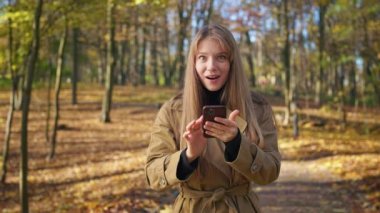 Front view of attractive, young lady standing in park, using smartphone. Glad, happy female reading, texting, wondering, looking at camera. Concept of modern, urban lifestyle.