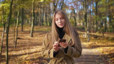 Front view of happy, cheerful lady walking in park in autumn. Pretty, young woman standing, using smartphone, looking at camera, smiling. Concept of harmony with nature.