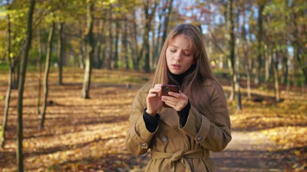 在阳光明媚的秋天里 一个年轻时髦的女人在公园里散步的侧视图 迷人的女士穿着时髦的衣服 使用智能手机 现代技术的概念 — 图库视频影像