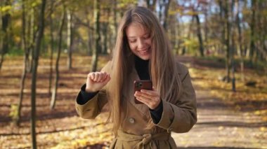 Front view of cheerful, positive lady walking, standing in park, dancing. Happy, glad, young female wearing stylish clothes, using smartphone. Concept of modern lifestyle.