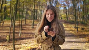 Parkta yürüyen, akıllı telefonlu, gülümseyen bir kadının ön görüntüsü. Şık, genç bayan akıllı telefon kullanıyor, mesajlaşıyor, mesajlaşıyor. Kentsel yaşam tarzı kavramı.