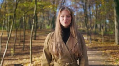 Front view of seductive, elegant lady walking in park, wearing fashionable clothes. Pretty, young female standing, looking at camera, waving. Concept of happiness and joy.
