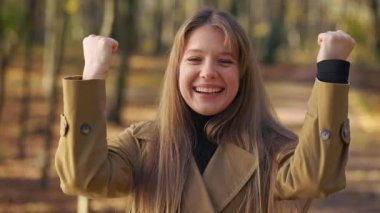 Front view of positive, cheerful lady walking, standing in park in autumn. Beautiful, happy woman looking at camera, showing hurray. Concept of happiness and sincere emotions.