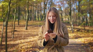 Side view of stylish, young female walking in park, using smartphone. Beautiful, cheerful lady typing, scrolling, messaging, reading, surprised. Concept of modern lifestyle.