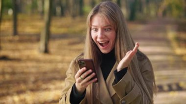 Side view of stylish, young female wearing fashionable clothes, walking in park. Attractive, happy lady standing, using smartphone, reading, wondering. Concept of sincere emotions.