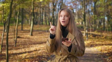 Front view of beautiful, young female standing in park, holding, using smartphone. Cheerful, creative lady texting, scrolling, messaging. Concept of modern technologies.
