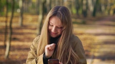 Front view of cheerful female standing, walking in park, dancing. Happy, glad lady using smartphone, texting, messaging, scrolling, smiling. Concept of happiness and joy.