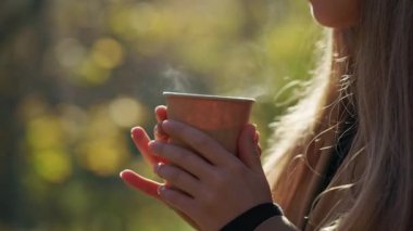 Close up of young lady walking, holding paper cup with coffee by hands. Attractive woman drinking hot coffee, smelling, enjoying aroma, smiling. Concept of happiness and joy.